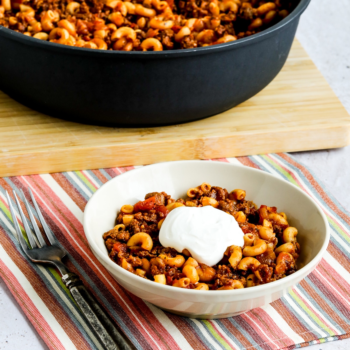 Square image for Better-Than-Mom's Hamburger Goulash with one serving shown in bowl with sour cream and pan of Goulash in back.