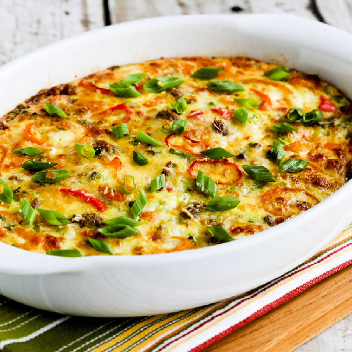 Square image of Breakfast Bake with Italian Sausage and Mini-Peppers shown in baking dish on striped napkin.