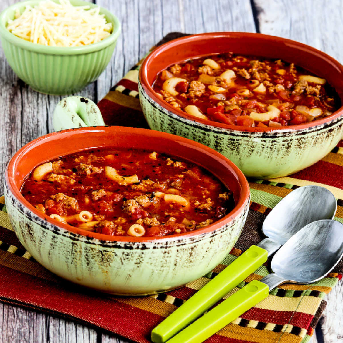 Square image for Italian Sausage, Tomato, and Pesto Soup with two bowls, two spoons, and Parmesan in the back.