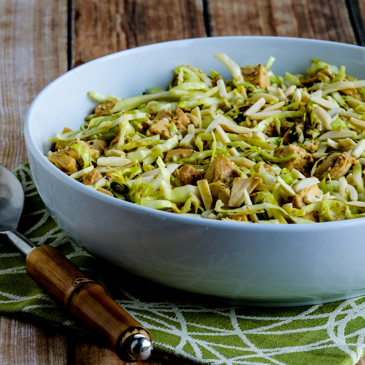 Square image of Sandee's Asian Chicken Cabbage Salad in bowl on green-white napkin with serving fork.