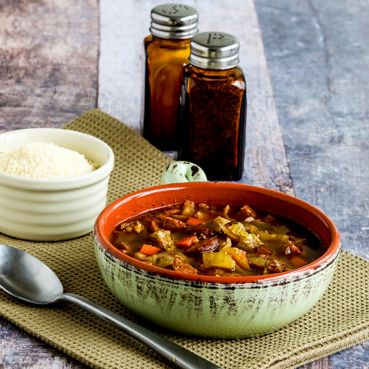 Square image for Sausage and Cabbage Soup shown in bowl with spoon, Parmesan, salt, and pepper.