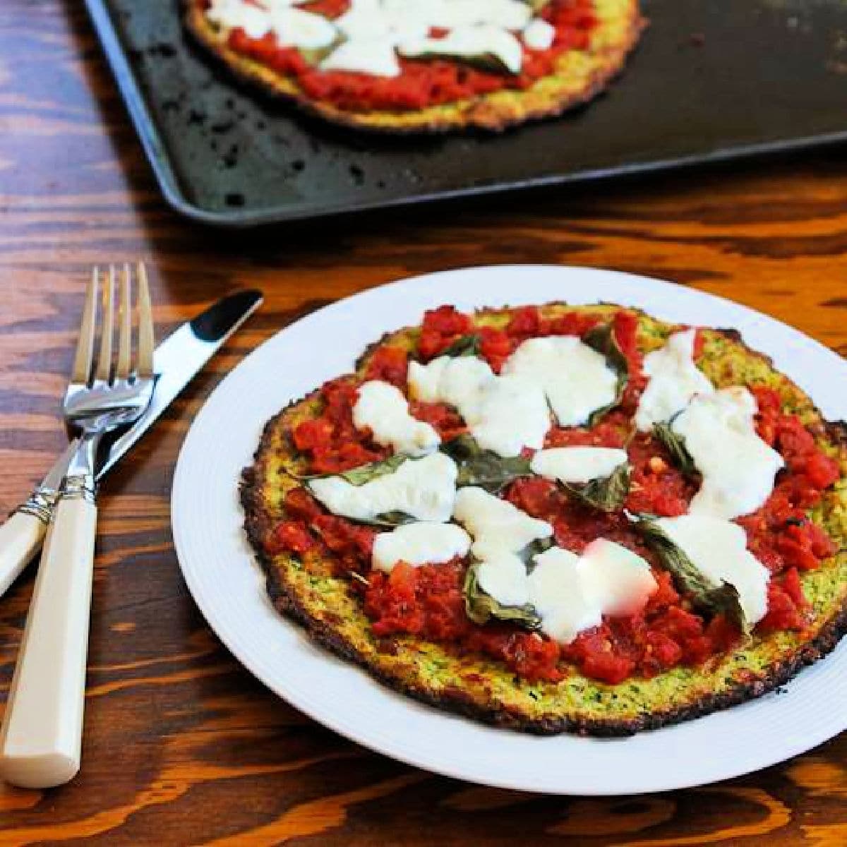 Square image of Zucchini Crust Pizza Margherita shown on serving plate with baking sheet in back.