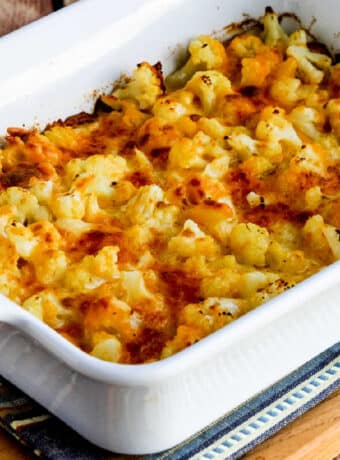 Square image of Easy Cheesy Baked Keto Cauliflower shown in baking dish on napkin and cutting board.