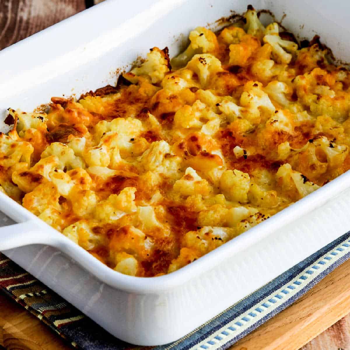 Square image of Easy Cheesy Baked Keto Cauliflower shown in baking dish on napkin and cutting board.