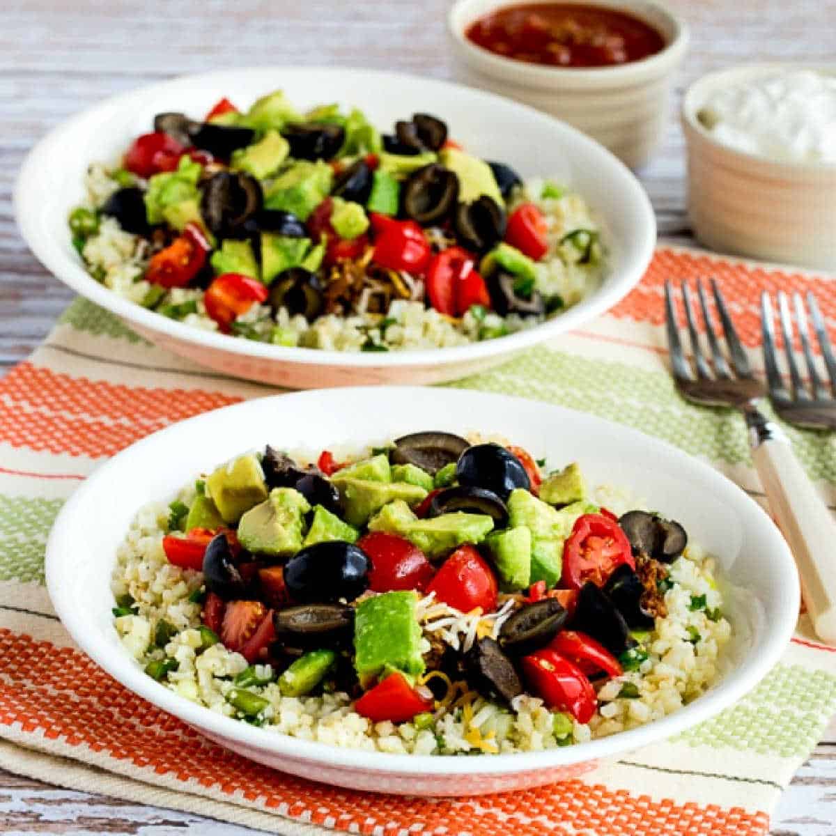 Square image of Ground Beef Cauliflower Rice Taco Bowls shown on napkin with forks and toppings in back.