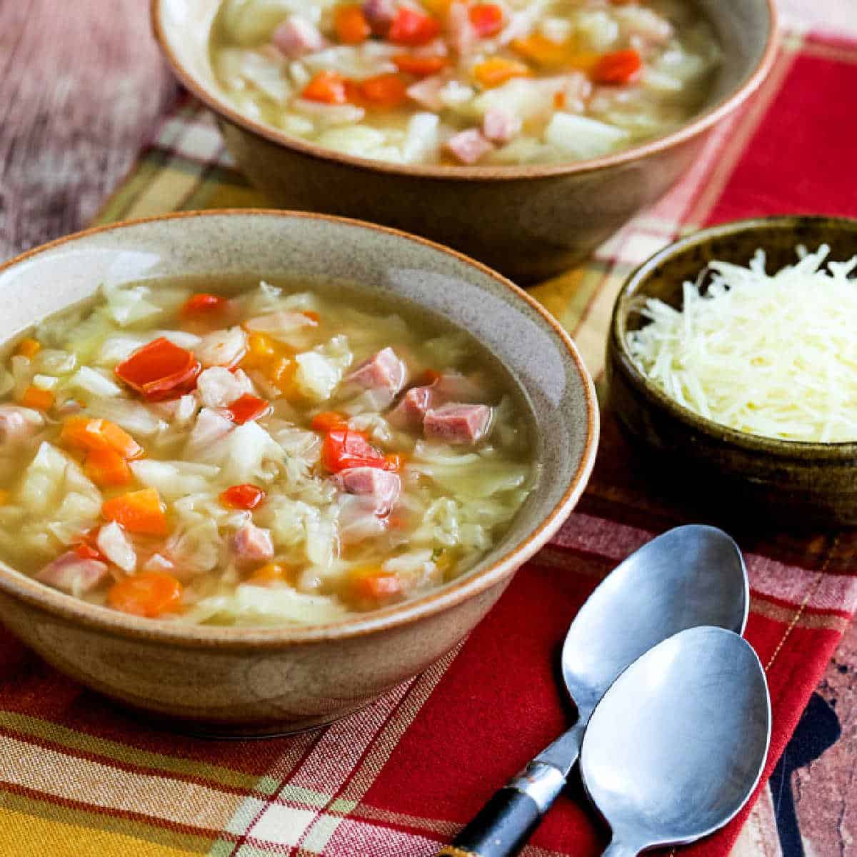 Square image of Instant Pot Ham and Cabbage Soup shown in two serving bowls with spoons and Parmesan Cheese.