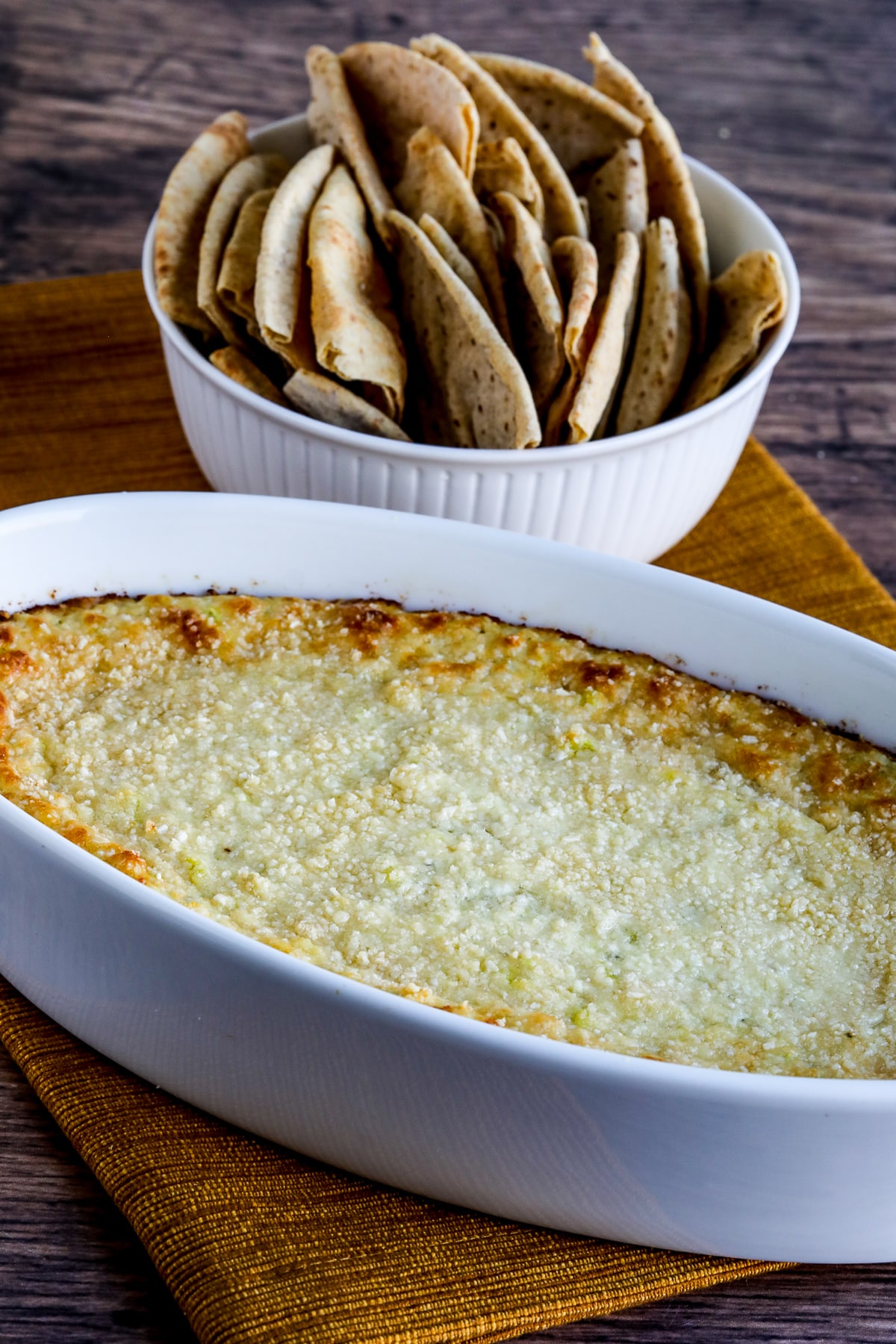 Hot Artichoke Dip with Peperoncini shown in baking dish with low-carb toasted pita bread in back.